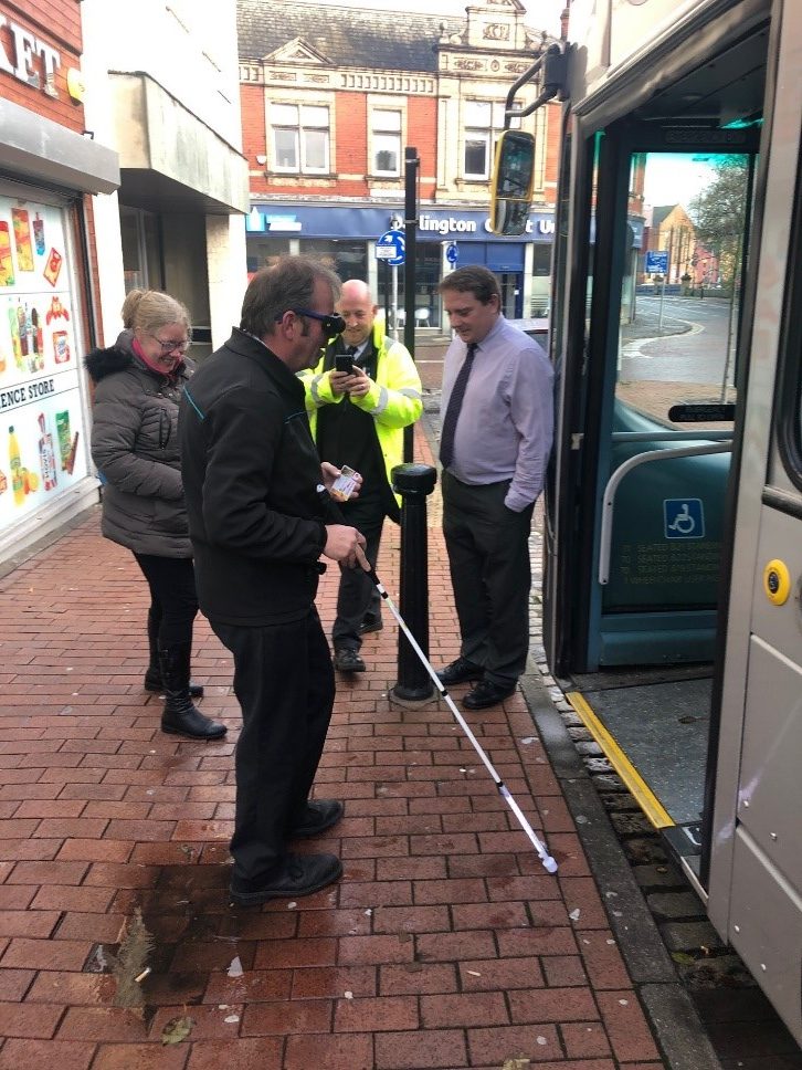 A man wears simulation goggles and uses a cane to board a bus